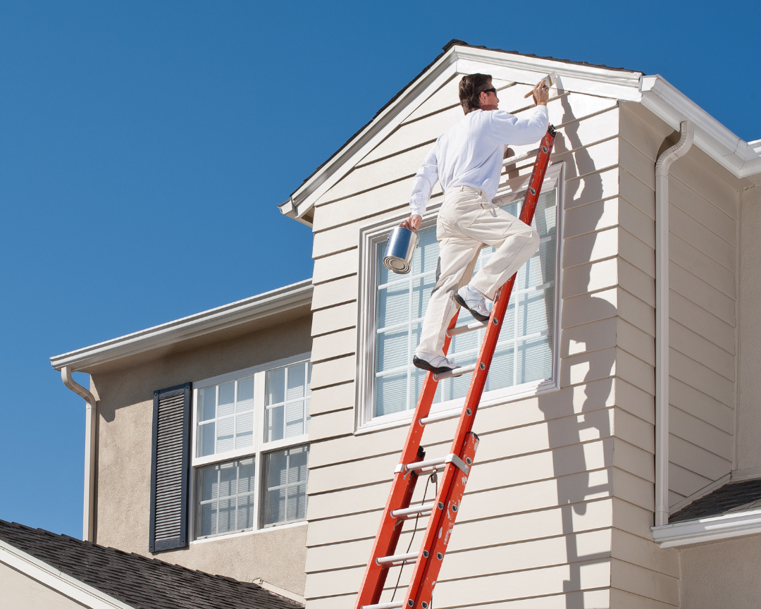 Person on a ladder painting the exterior of a house against a clear blue sky.